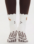 Back view of both feet wearing the Corgi grip socks in oat milk with scalloped cuffs, embroidered corgi design, and brown paw print grip soles with Almla wordmark