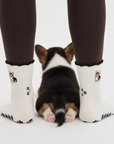 Real tri-color corgi puppy sitting between two feet wearing the Tri-Color Corgi grip socks with black cuffs, embroidered corgi design, and black paw print grip soles