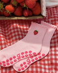 Pink strawberry cute pilates socks laid on a red gingham cloth next to a box of fresh strawberries.