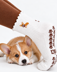 Corgi puppy lying down with its head resting under a foot wearing the Corgi grip sock in oat milk with ruffle cuff, embroidered corgi design, and brown paw print grip sole