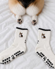 A real corgi lying on a bed next to a pair of The Tri Color Corgi Grip Sock, showing the embroidered corgi design and paw-print grip soles