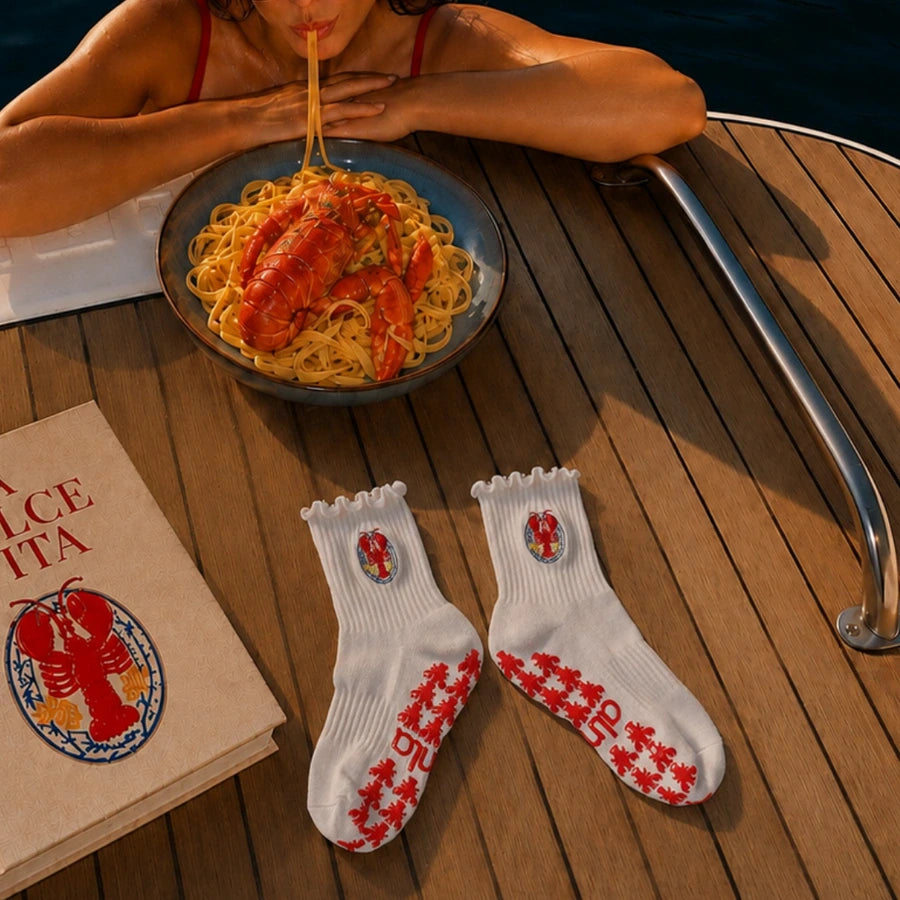 Pair of white lobster-crest sticky socks for barre class on a boat deck beside lobster pasta and a book.