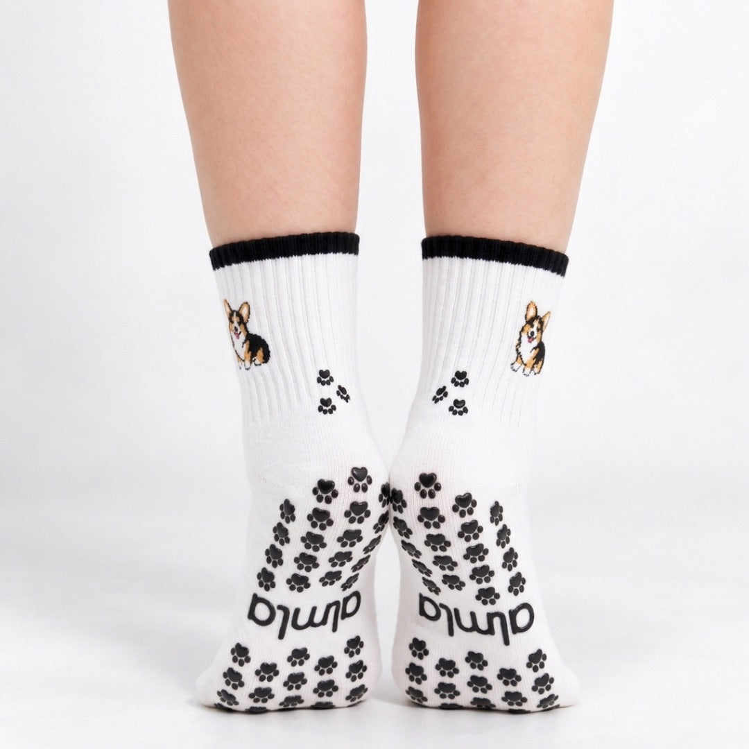 Model standing in a pair of The Tri Color Corgi Grip Sock, front view showing the embroidered tri-color corgi on each ankle and paw-print grip soles with the Almla logo