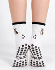 Model standing in a pair of The Tri Color Corgi Grip Sock, front view showing the embroidered tri-color corgi on each ankle and paw-print grip soles with the Almla logo