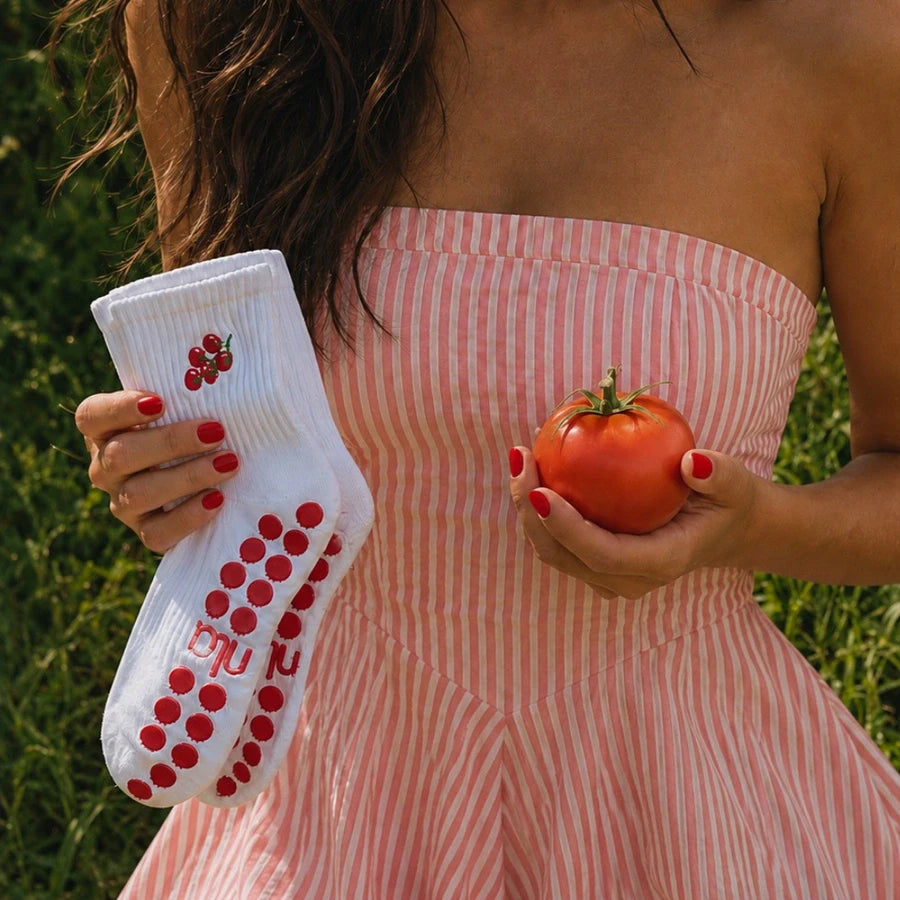 Woman in a pink striped dress holding a white cherry-tomato pilates grip sock and a ripe tomato outdoors.