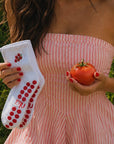 Woman in a pink striped dress holding a white cherry-tomato pilates grip sock and a ripe tomato outdoors.