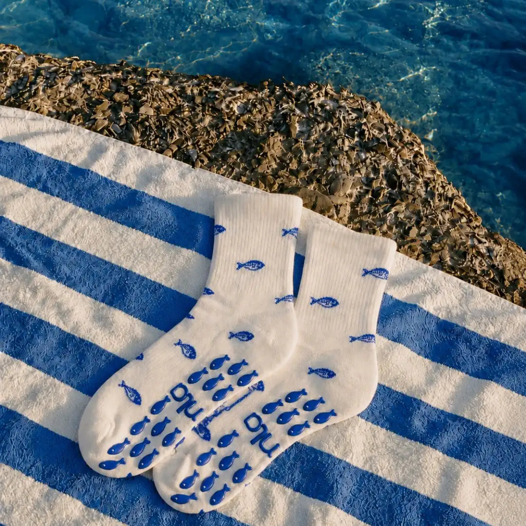 Pair of white fish-print cute pilates socks laid on a blue striped towel by the sea with blue fish grip soles.