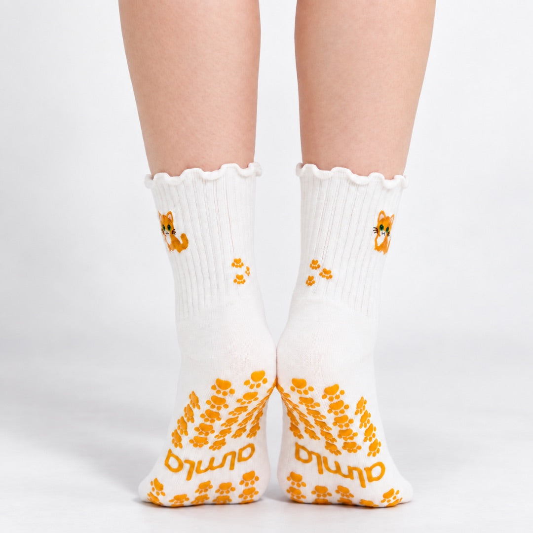 Model standing in a pair of The Orange Cat Grip Sock, front view showing the embroidered orange tabby cat on each ankle and paw-print grip soles with the Almla logo