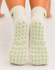 Back view of raised feet wearing cream margarita yoga socks with grips for women and pale green grip soles.
