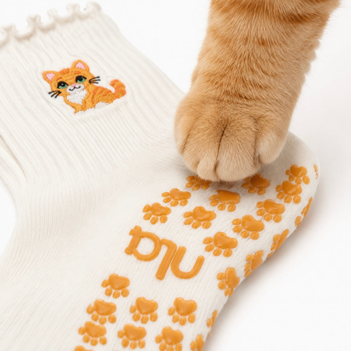 A real ginger cat's paw resting on the paw-print grip sole of The Orange Cat Grip Sock, showing the embroidered orange tabby cat design