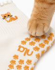 A real ginger cat's paw resting on the paw-print grip sole of The Orange Cat Grip Sock, showing the embroidered orange tabby cat design