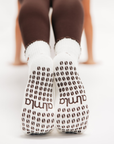 Back view of raised feet in brown leggings wearing white non skid socks for pilates with brown coffee-bean grips.