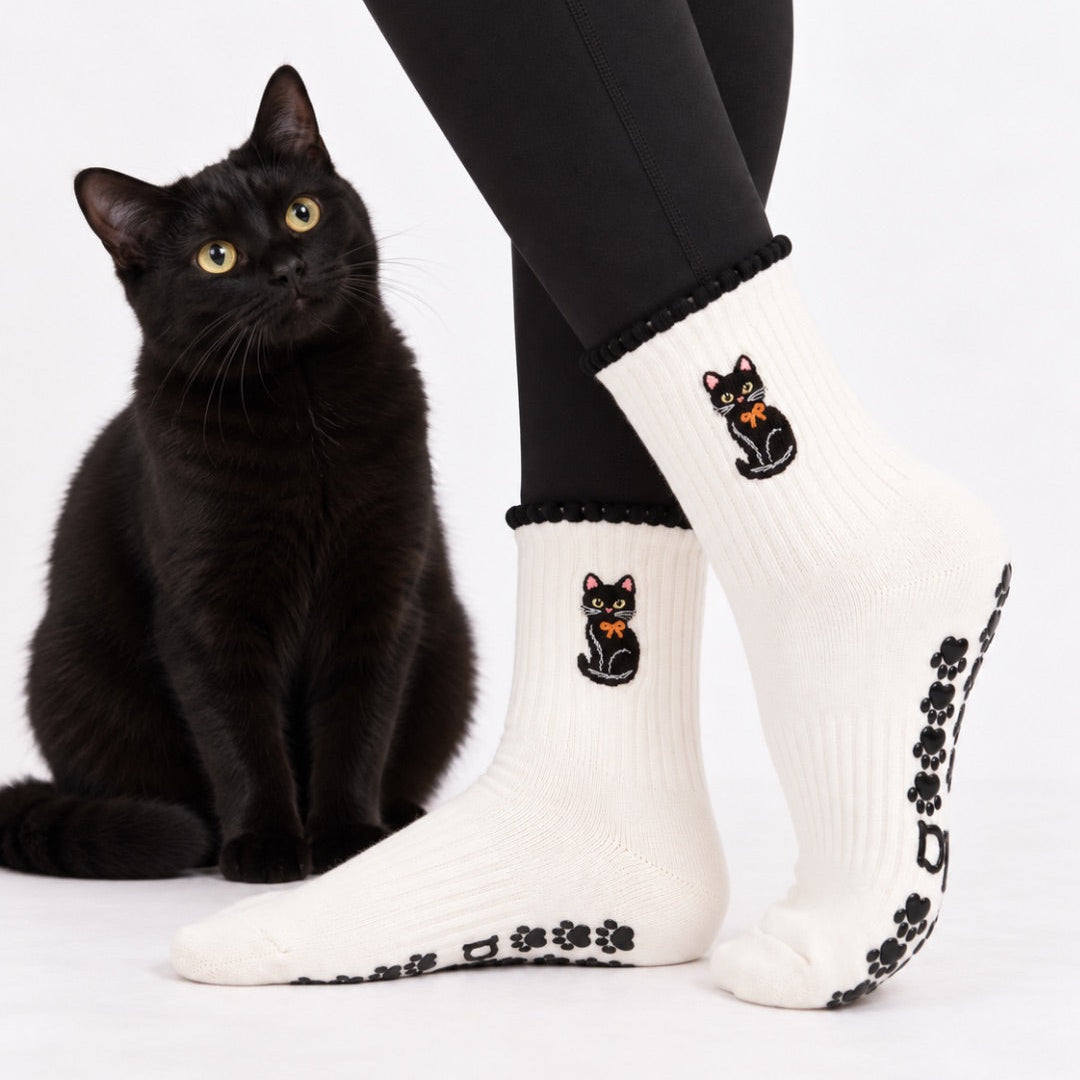 Black cat sitting next to a model wearing the Black Cat Grip Sock with embroidered cat and orange bow