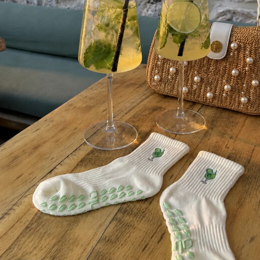 Pair of white margarita best yoga socks with grips on a table beside two mojito cocktails and a pearl bag.