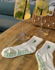 Pair of white margarita best yoga socks with grips on a table beside two mojito cocktails and a pearl bag.