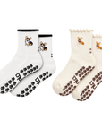 The Corgi Obsessed Duo grip socks flat lay: Tri-Color Corgi sock with black cuff and black paw print grip sole next to the Corgi sock with scalloped cuff and paw print grip sole, both on an oat milk base with embroidered corgi design