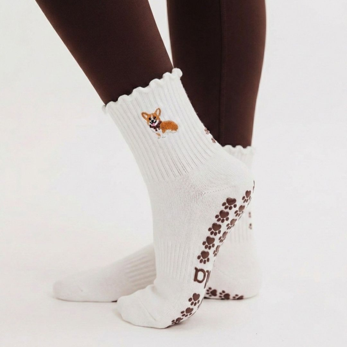 Person standing wearing the Corgi grip sock in oat milk with scalloped cuff, embroidered corgi design, and brown paw print grip sole