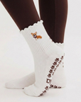 Person standing wearing the Corgi grip sock in oat milk with scalloped cuff, embroidered corgi design, and brown paw print grip sole