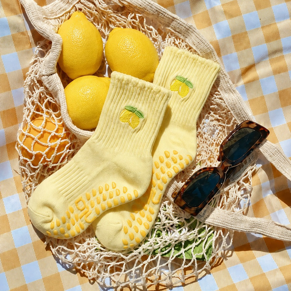 Pair of yellow lemon socks with grips for pilates resting in a mesh bag with lemons and sunglasses.