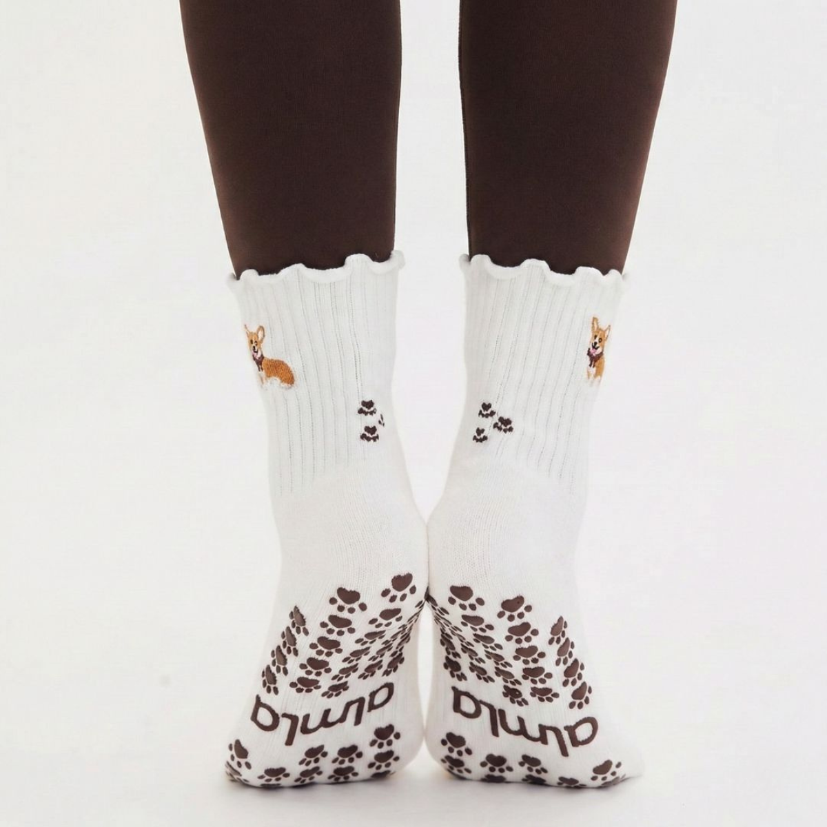 Back view of both feet wearing the Corgi grip socks in oat milk with scalloped cuffs, embroidered corgi design, and brown paw print grip soles with Almla wordmark