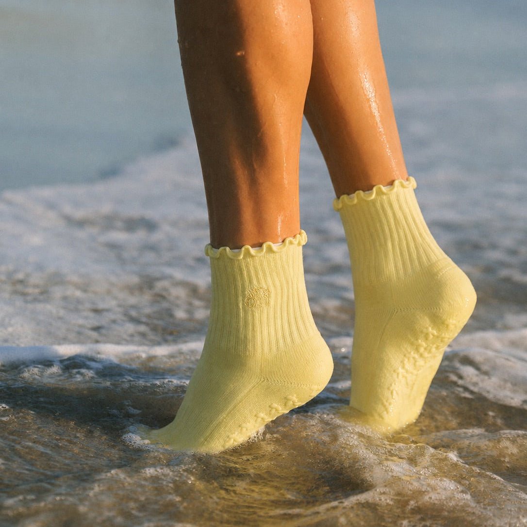 Butter yellow grip socks worn ankle-deep in ocean water at the shoreline