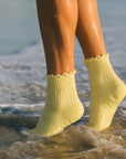 Butter yellow grip socks worn ankle-deep in ocean water at the shoreline