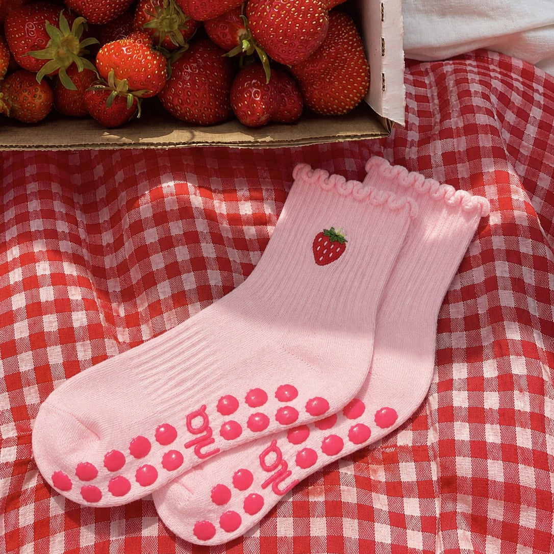 Pink strawberry cute pilates socks laid on a red gingham cloth next to a box of fresh strawberries.