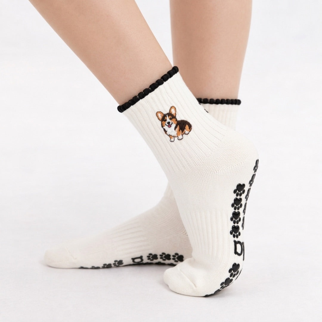 Model wearing The Tri Color Corgi Grip Sock, crossed feet showing the paw-print grip soles and embroidered tri-color corgi on the cuff