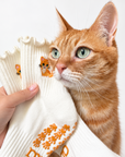 A real orange tabby cat nuzzling a hand holding The Orange Cat Grip Sock, showing the embroidered cat design and paw-print grip sole