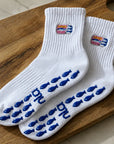 White sardine-tin embroidered barre grip socks with blue fish-shaped grips arranged on a wooden board.
