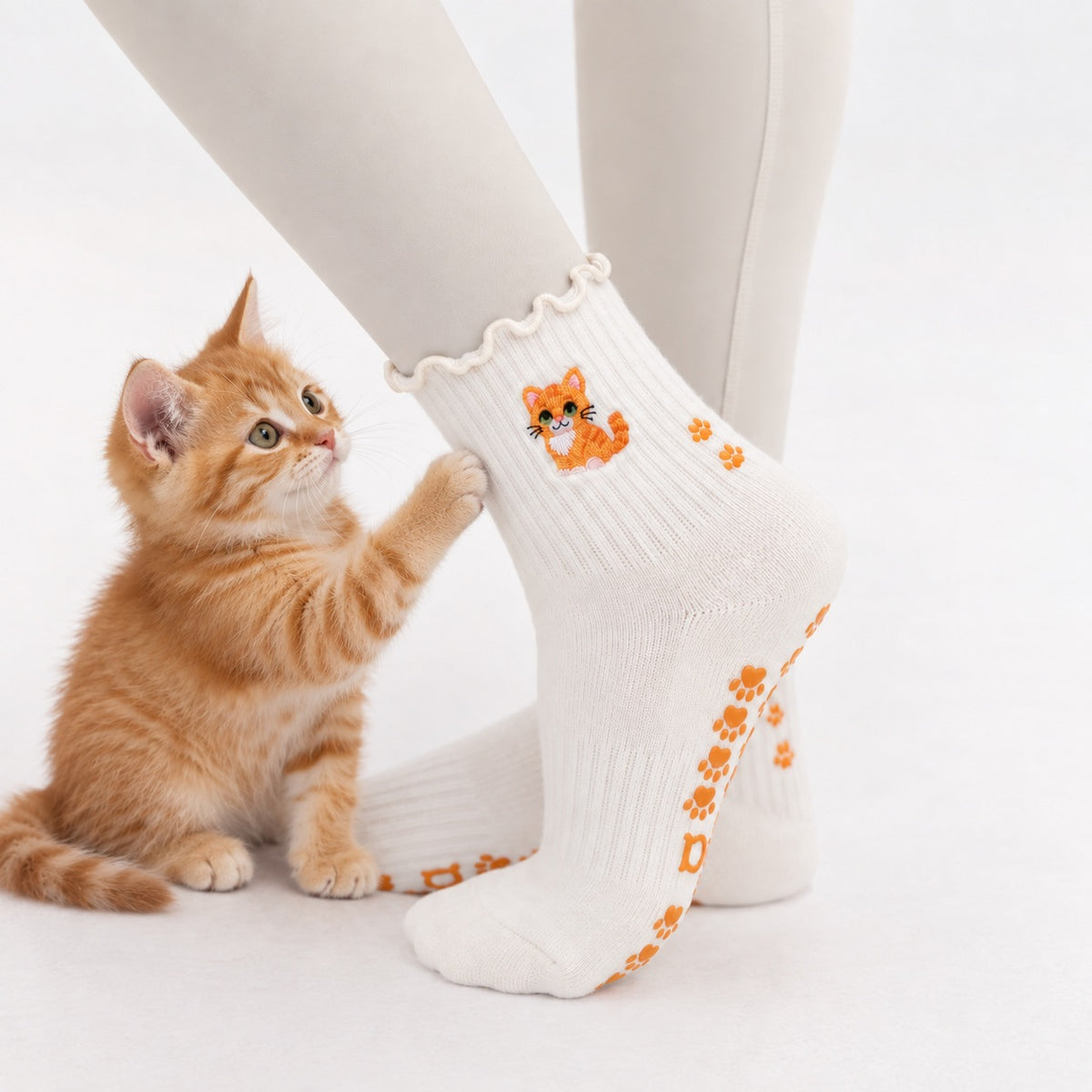 A ginger kitten standing on hind legs against a model's leg wearing The Orange Cat Grip Sock, showing the embroidered orange tabby cat design and paw-print grip soles