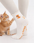 A ginger kitten standing on hind legs against a model's leg wearing The Orange Cat Grip Sock, showing the embroidered orange tabby cat design and paw-print grip soles
